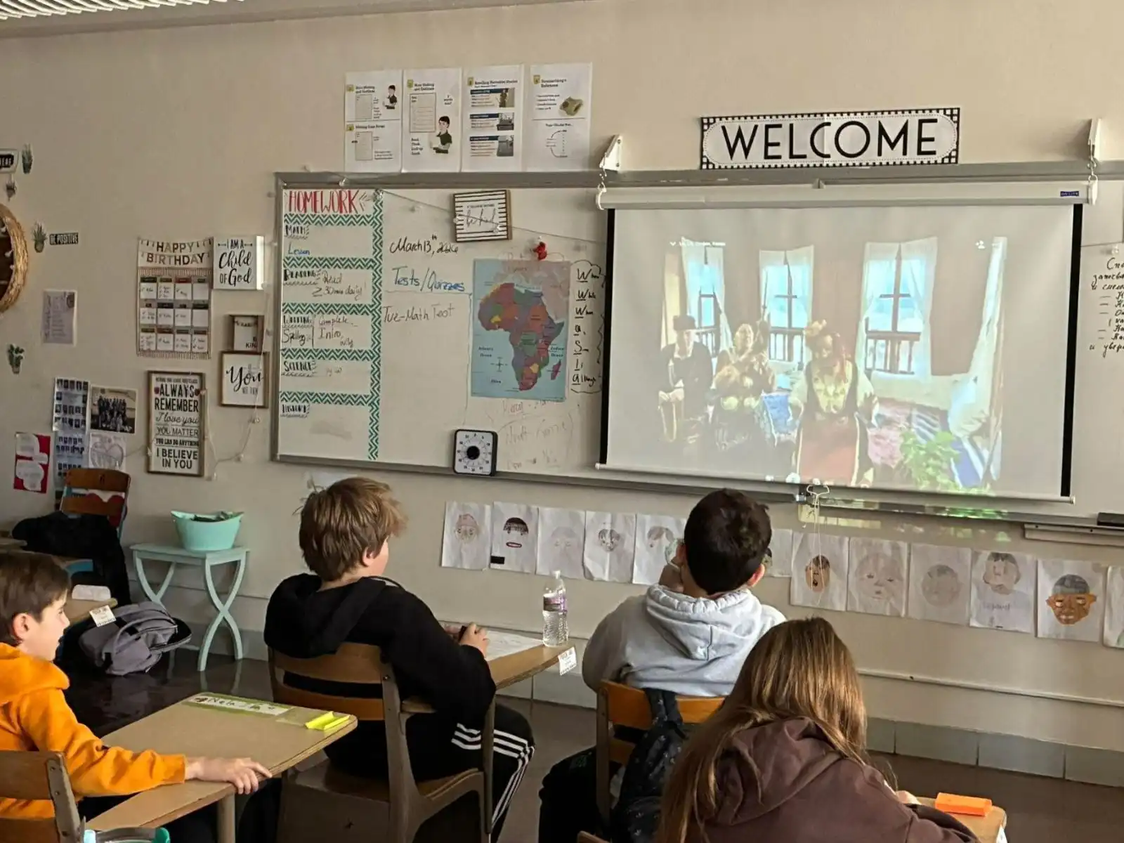 Students attentively watch a presentation on a screen in a classroom decorated with educational posters and a "WELCOME" sign.