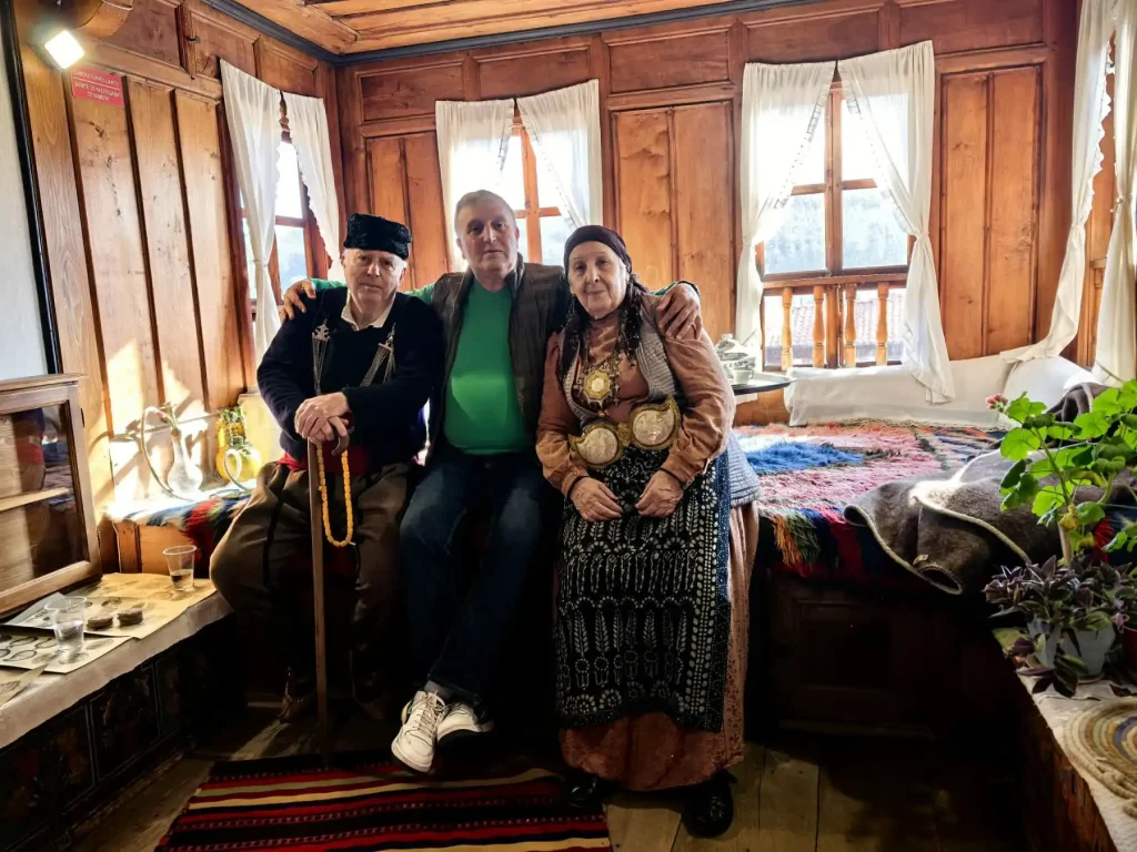 Three individuals sit together in a rustic wooden room, adorned with traditional Bulgarian textiles. Warm sunlight filters through the windows.
