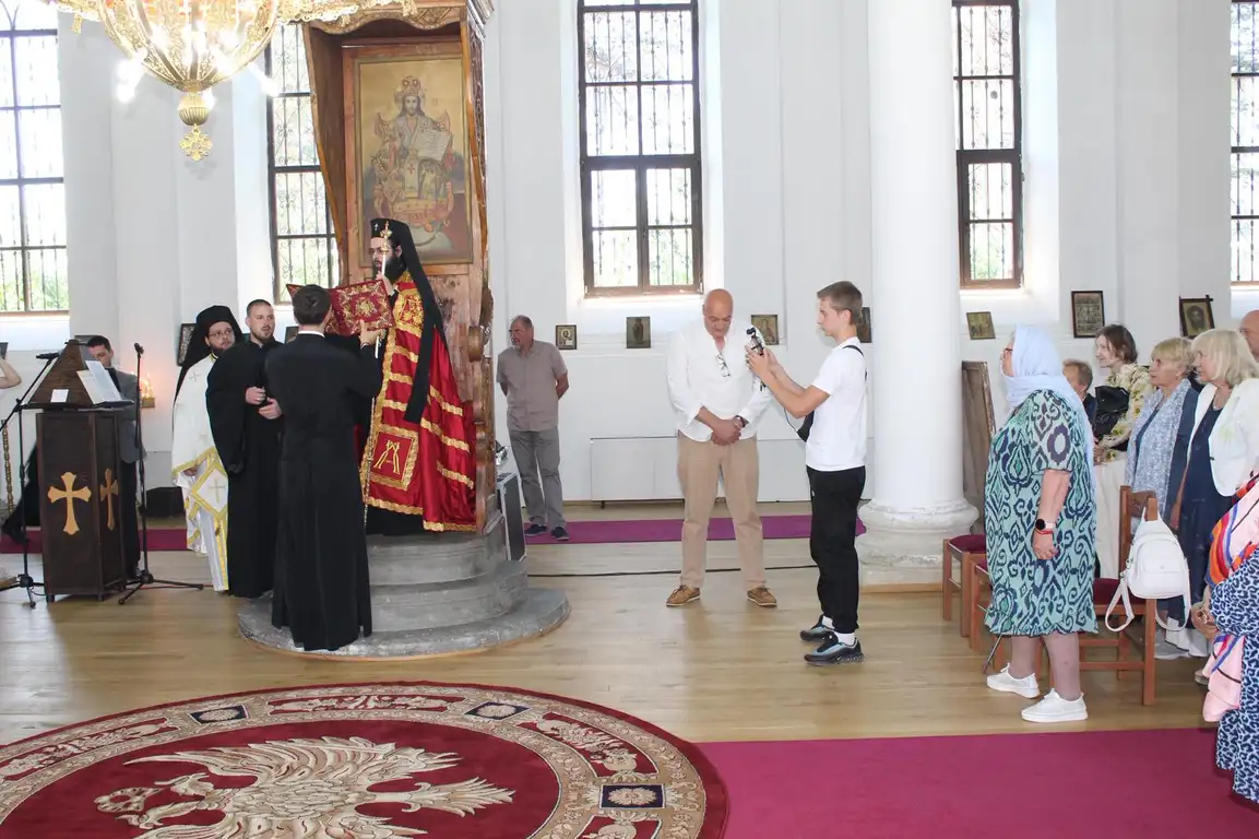 Събитие в църква с монаси и миряни, облечени в традиционни одежди, на фона на икона и декорации.