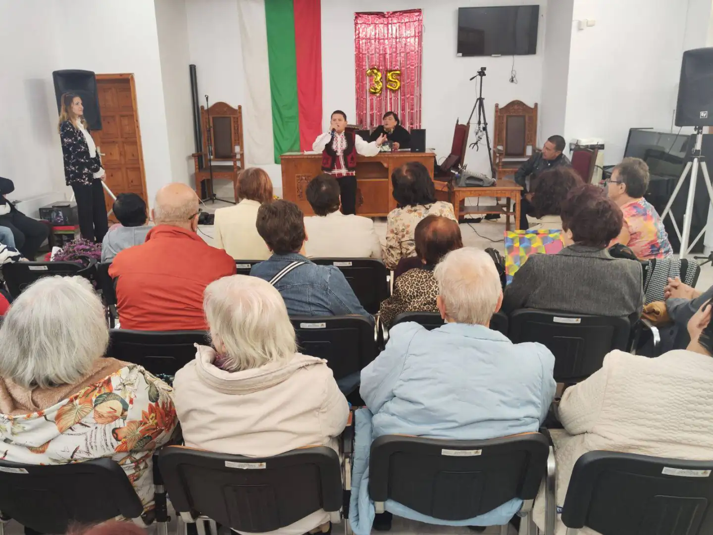 Събитие за възрастни хора с говорители и публика в зала с българския флаг на заден план.