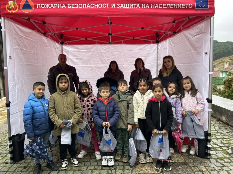 Деца заедно с възрастни под шатра на пожарната служба, получаващи подаръци и информация за безопасност.