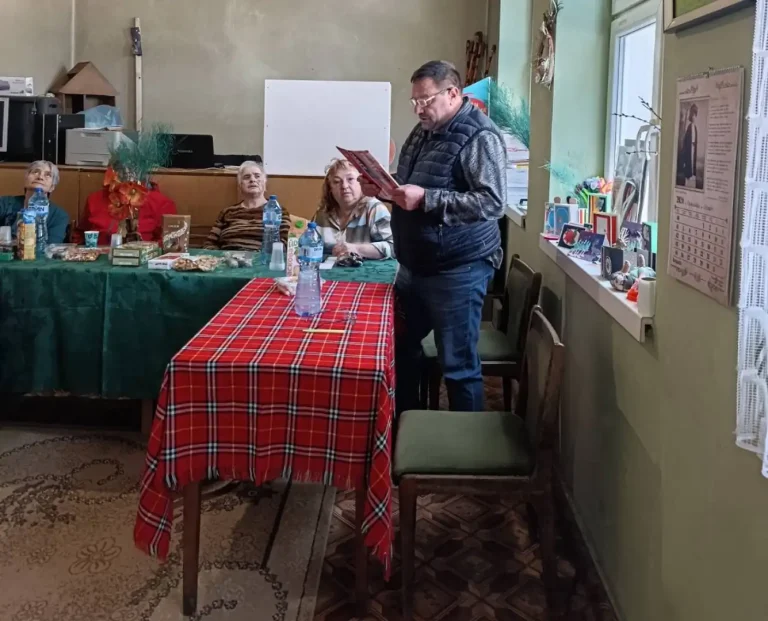 Събитие с възрастни хора, където мъж чете пред група, облечени в комфортна обстановка.