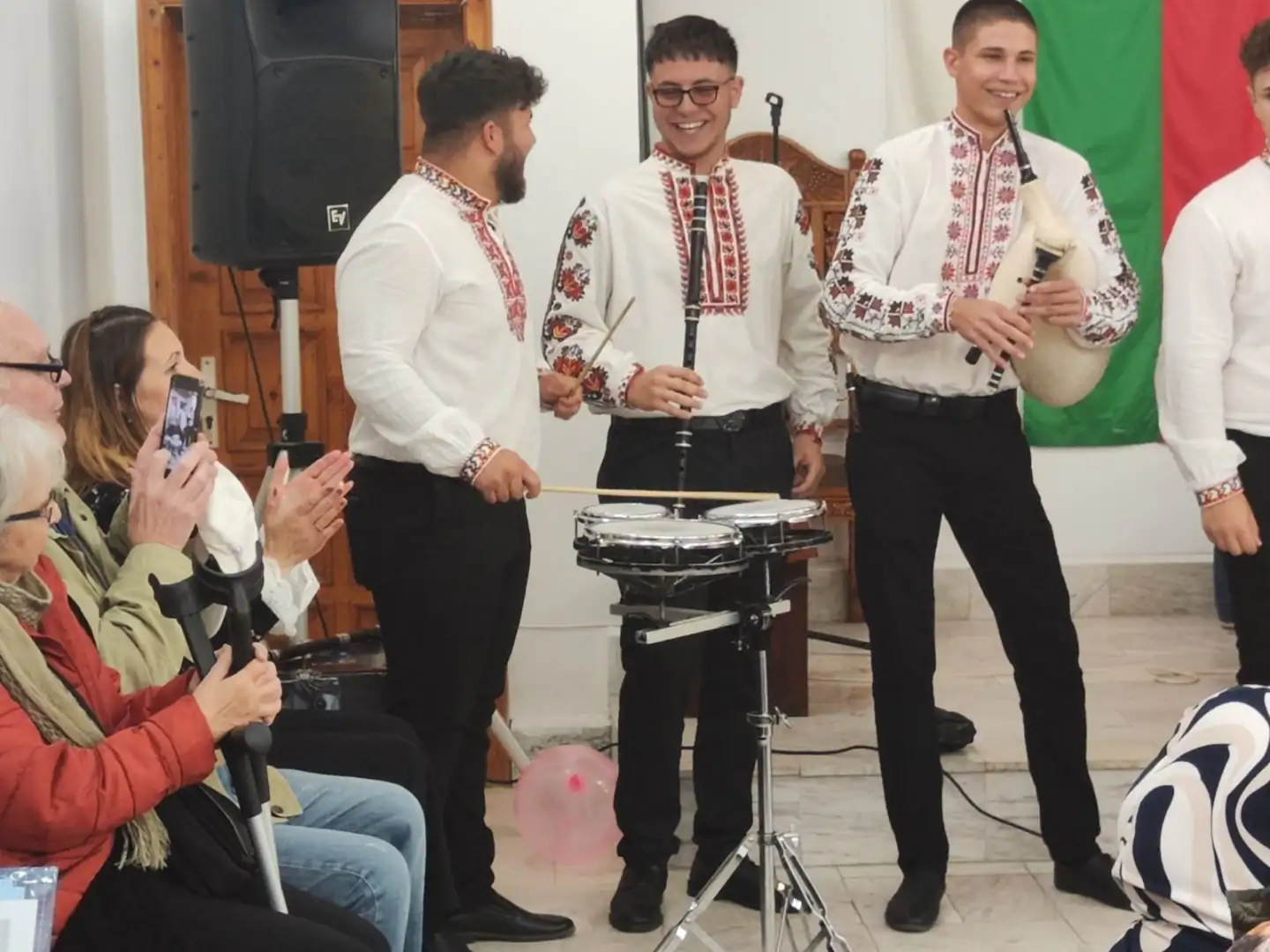 Музикална група в традиционни носии, представяща български фолклор на сцената с ентусиазирана публика.